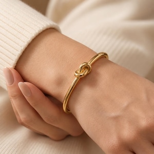 Può includere: Un bracciale rigido dorato con un design a nodo. Questo semplice ed elegante gioiello presenta un nodo come punto focale, aggiungendo un tocco di raffinatezza. Il bracciale è indossato su un polso.