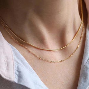 May include: A layered gold-tone necklace set consisting of two distinct chains. The upper chain is a smooth, flexible snake chain, while the lower chain is a delicate cable chain featuring small, evenly spaced gold beads along its length.