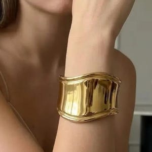 May include: A wide, polished gold-tone cuff bracelet featuring a smooth, undulating wave-like surface. The metal has a high-shine, reflective finish that catches the light, creating a fluid, organic shape that wraps around the wrist area.