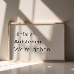 Puede incluir: Un cuadro rectangular con marco de madera clara que presenta el texto en alemán "Hinfallen. Aufstehen. Weitergehen." en una fuente sans-serif gris oscuro sobre un fondo color crema, iluminado por sombras de luz natural.