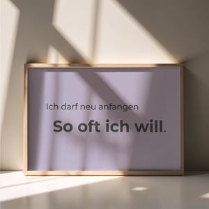 Könnte beinhalten: Ein gerahmter Druck mit helllila Hintergrund und dem deutschen Text "Ich darf neu anfangen So oft ich will." Der Rahmen ist hellbraun, und der Druck wird auf einer weißen Oberfläche angezeigt.