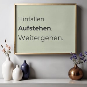 Puede incluir: Una pieza de arte mural rectangular en un marco dorado fino con un fondo verde salvia claro y el texto negro "Hinfallen. Aufstehen. Weitergehen." en tres líneas. La obra de arte se encuentra sobre tres jarrones.