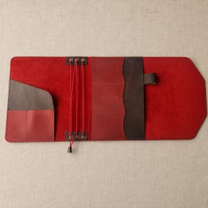 May include: A flat, open leather journal cover in vibrant red with dark brown leather pockets and accents. The interior features three vertical elastic cords for securing inserts, multiple card slots, and a pen loop on the right side.