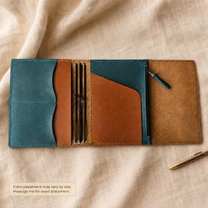 May include: A leather notebook cover featuring a color-blocked design in teal and tan. The interior includes multiple pockets, a zippered compartment with a metal pull, and elastic bands for securing inserts. Text on the image reads: Color placement may vary by size. Message me for exact placement.