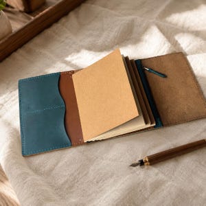 Pode incluir: Uma capa de couro para caderno com um bolso azul-petr&oacute;leo &agrave; esquerda, interior castanho e um compartimento com fecho &agrave; direita. O caderno cont&eacute;m v&aacute;rias p&aacute;ginas de papel castanho claro e uma caneta de tinta permanente castanha.