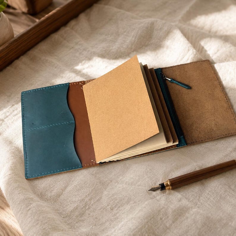 Pode incluir: Uma capa de couro para caderno com um bolso azul-petr&oacute;leo &agrave; esquerda, interior castanho e um compartimento com fecho &agrave; direita. O caderno cont&eacute;m v&aacute;rias p&aacute;ginas de papel castanho claro e uma caneta de tinta permanente castanha.