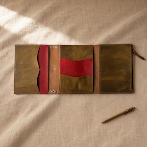 May include: A tri-fold leather wallet in olive green with contrasting red interior pockets and card slots. The wallet features a zipped compartment on the right side and metal rivets along the central spine, accompanied by a slim gold-tone pen.