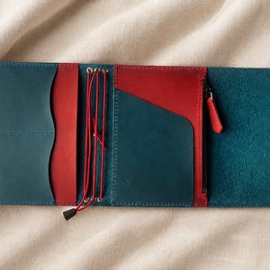 Pode incluir: Uma capa de caderno em couro multicolorida com exterior e pain&eacute;is internos em azul-petr&oacute;leo e bolsos de couro vermelho contrastantes. A capa inclui um sistema de cord&atilde;o el&aacute;stico vermelho vertical para fixar encartes e um bolso lateral com z&iacute;per.