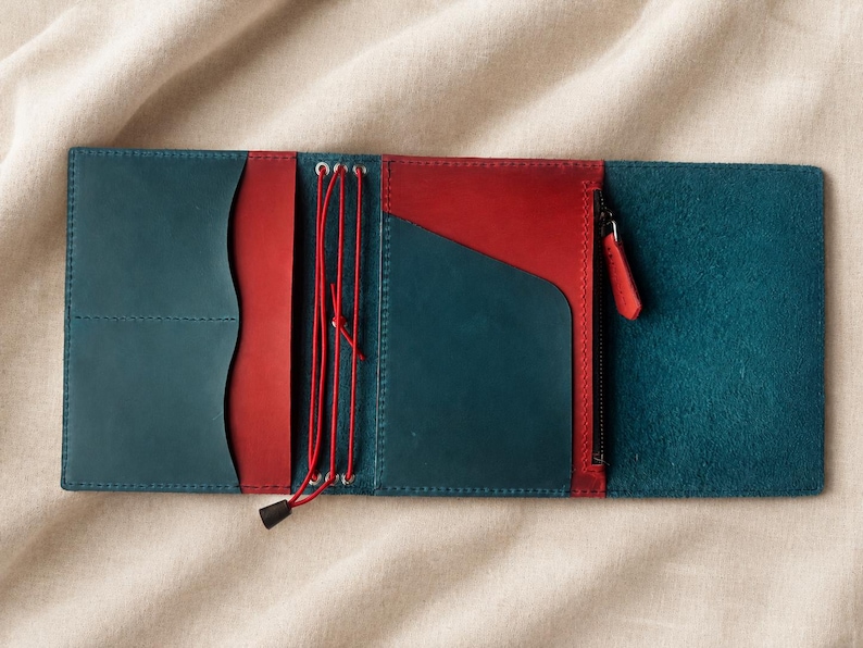 Pode incluir: Uma capa de caderno em couro multicolorida com exterior e pain&eacute;is internos em azul-petr&oacute;leo e bolsos de couro vermelho contrastantes. A capa inclui um sistema de cord&atilde;o el&aacute;stico vermelho vertical para fixar encartes e um bolso lateral com z&iacute;per.