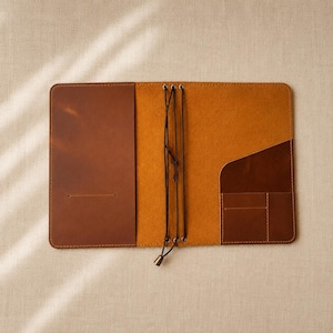 May include: A tan leather notebook cover featuring a smooth exterior finish and a textured interior. The interior includes a vertical pocket on the left, a diagonal pocket on the right, and a central elastic cord system with metal eyelets.
