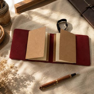 May include: A deep red suede leather notebook cover featuring two internal slots holding separate paper inserts with light brown covers. A black elastic band is attached to the top edge of the cover. A brown and gold fountain pen lies nearby.