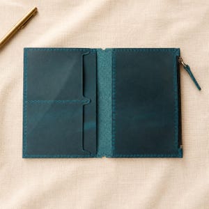 May include: A teal leather passport holder or wallet featuring two card slots on the left side and a zippered compartment on the right side. The interior is constructed with visible stitching along the edges and a matching teal leather pull tab.
