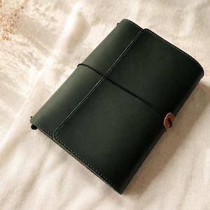 Pode incluir: Uma capa de di&aacute;rio em couro verde-escuro com acabamento mate liso, presa por uma fina fita el&aacute;stica preta que envolve verticalmente o centro. Uma pequena aba de couro castanho claro est&aacute; presa ao el&aacute;stico no lado direito da capa.