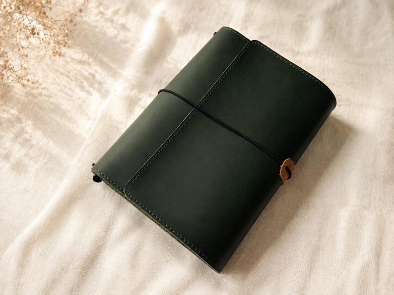 Pode incluir: Uma capa de di&aacute;rio em couro verde-escuro com acabamento mate liso, presa por uma fina fita el&aacute;stica preta que envolve verticalmente o centro. Uma pequena aba de couro castanho claro est&aacute; presa ao el&aacute;stico no lado direito da capa.