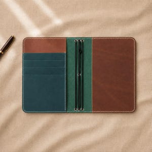 May include: A multi-coloured leather notebook cover featuring a brown exterior, a teal interior with four horizontal card slots, and a green centre spine section with two black elastic bands for securing inserts, accompanied by a black fountain pen.