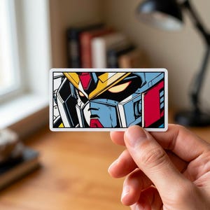 Puede incluir: Una pegatina rectangular con una ilustraci&oacute;n estilizada de la cara de un robot. El robot tiene un casco amarillo, detalles en rojo y detalles en azul y blanco. Los ojos del robot brillan en naranja sobre un fondo negro.