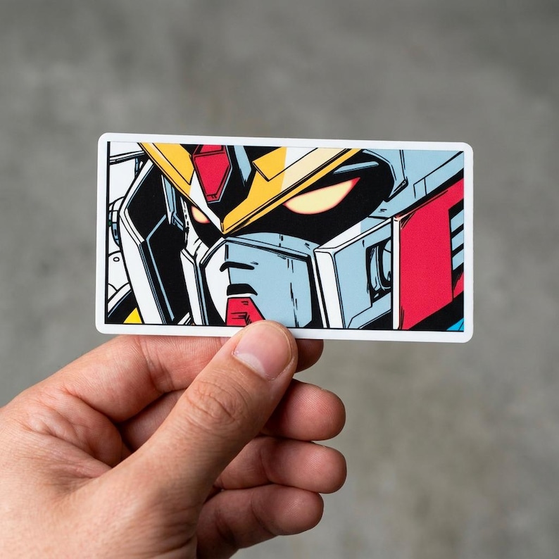 Puede incluir: Una pegatina rectangular que presenta un primer plano de la cara de un robot. El robot tiene un casco amarillo, detalles en rojo y una armadura azul y blanca. Los ojos del robot brillan en rojo sobre un fondo negro. La pegatina est&aacute; sujeta por una mano.