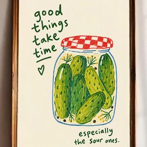 Op de afbeelding: Een ingelijste kunstdruk met een handgetekende illustratie van een glazen pot gevuld met groene augurken. De pot heeft een rood-wit geblokt deksel. De tekst luidt: good things take time, especially the sour ones, met een klein hartje erbij.