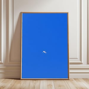 Op de afbeelding: Een ingelijste kunstdruk met een helderblauwe lucht en een kleine, witte figuur die in het midden zweeft. Het kunstwerk wordt gepresenteerd in een houten lijst, leunend tegen een witte muur en een houten vloer.