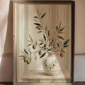 Può includere: Una stampa artistica da parete rettangolare incorniciata raffigurante un vaso in ceramica dipinto con un ramo di ulivo all'interno. Il vaso poggia su un tavolo di legno su uno sfondo a strisce verticali verde chiaro e crema. La cornice è argentata.