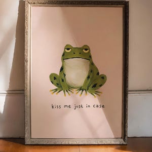Op de afbeelding: Ingelijste kunstdruk met een groene kikker met zwarte vlekken en een witte buik op een roze achtergrond. De kikker heeft een chagrijnige uitdrukking. De tekst "kiss me just in case" staat eronder.