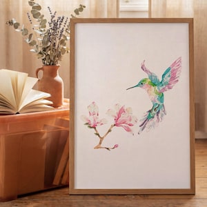 Op de afbeelding: Een ingelijste aquarelprint met een kolibrie en magnoliabloemen. De kolibrie heeft groene, blauwe en roze tinten. De lijst is van lichtbruin hout. De print wordt op een houten oppervlak gepresenteerd.