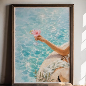 May include: A framed art print depicting a person holding a pink cocktail with a cherry and lemon twist, positioned by a turquoise swimming pool. The scene includes a straw hat, a patterned fabric, and gold bangles, all set in a dark wood frame.
