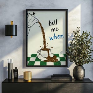 Op de afbeelding: Ingelijste kunstafdruk met een koffiepot die koffie in een kopje giet, met de woorden "tell me when" in zwart en blauw. De achtergrond heeft een groen-wit geblokt patroon. Het kunstwerk wordt tentoongesteld op een zwarte kast.