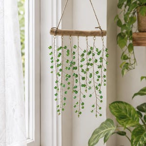 May include: A hanging wall decoration featuring a horizontal piece of driftwood suspended by a twine cord and a metal hook. Seven strands of thin wire hang from the wood, each adorned with small, translucent green beads resembling trailing vines.