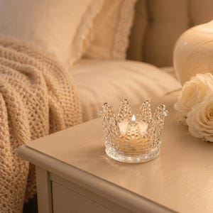 Peut inclure: Un bougeoir en verre transparent en forme de couronne avec une surface texturée et facettée. Le bougeoir contient une petite bougie chauffe-plat ronde avec une mèche allumée, diffusant une lueur chaude depuis le centre de la structure.