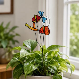 May include: A stained glass plant stake featuring a red and orange flower, a yellow leaf, and a blue dragonfly. The decorative piece is supported by a thin black metal rod and is inserted into a green leafy plant housed in a light grey pot.