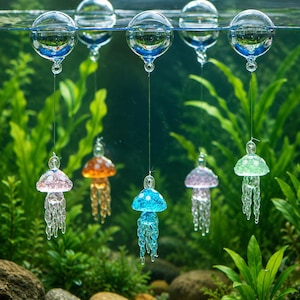 May include: Five glass jellyfish ornaments suspended by thin lines from clear glass spheres floating on the water surface. Each jellyfish features a distinct color including pink, orange, blue, purple, and green with white spots and dangling tentacles.