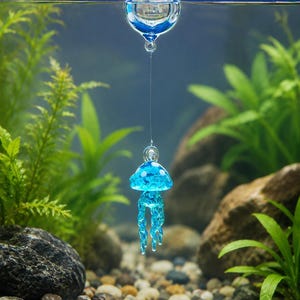 May include: A clear glass sphere floating at the water surface, connected by a thin transparent line to a small, translucent blue glass jellyfish ornament with white spots and dangling tentacles suspended underwater in an aquarium.