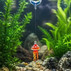 May include: A clear glass sphere floats on the water surface, connected by a thin line to a small, red, mushroom-shaped glass ornament with white spots and dangling orange tentacles, suspended underwater in an aquarium setting.