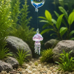 May include: A clear glass sphere floating at the water surface, connected by a thin filament to a small, iridescent pink and white glass jellyfish ornament with twisted tentacles suspended underwater in an aquarium setting.