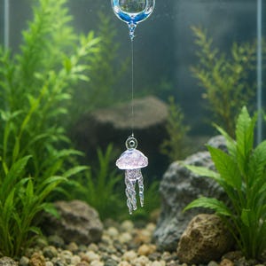 May include: A clear glass jellyfish ornament suspended by a thin filament from a floating glass sphere. The jellyfish has a translucent pinkish-purple bell and elongated, twisted tentacles, hanging vertically within a clear liquid environment.