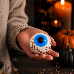 May include: A spherical glass ornament designed to resemble a human eyeball, featuring a bright blue iris, a solid black pupil, and intricate red vein details set against a white background, crafted with a smooth, glossy finish.