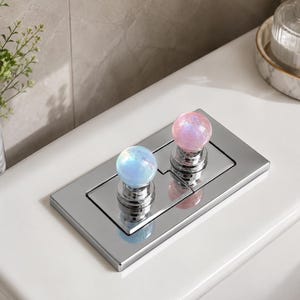 May include: A rectangular chrome toilet flush plate featuring two circular buttons. The left button is a light blue sphere with iridescent flecks, and the right button is a light pink sphere with iridescent flecks, both mounted on chrome bases.