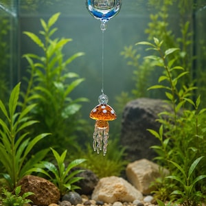 May include: A clear glass sphere floats on the water surface, connected by a thin transparent line to a small orange glass mushroom ornament with white spots and dangling clear glass tentacles suspended in the water.