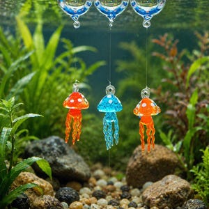 May include: Three glass jellyfish ornaments suspended by thin lines from three clear glass spheres floating on the water surface. The jellyfish are colored orange, blue, and orange, each featuring white spots on their caps and dangling tentacles.