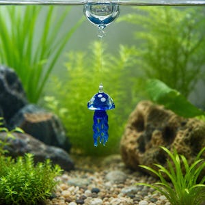 May include: A blue glass jellyfish ornament with white spots suspended from a clear glass sphere floating at the water surface. The jellyfish has a rounded bell top and multiple dangling, wavy tentacles hanging vertically in the water.