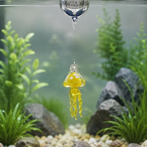 May include: A clear glass sphere floats at the water surface, connected by a thin filament to a small, yellow glass jellyfish ornament. The jellyfish features a rounded bell with white spots and several dangling, twisted glass tentacles.