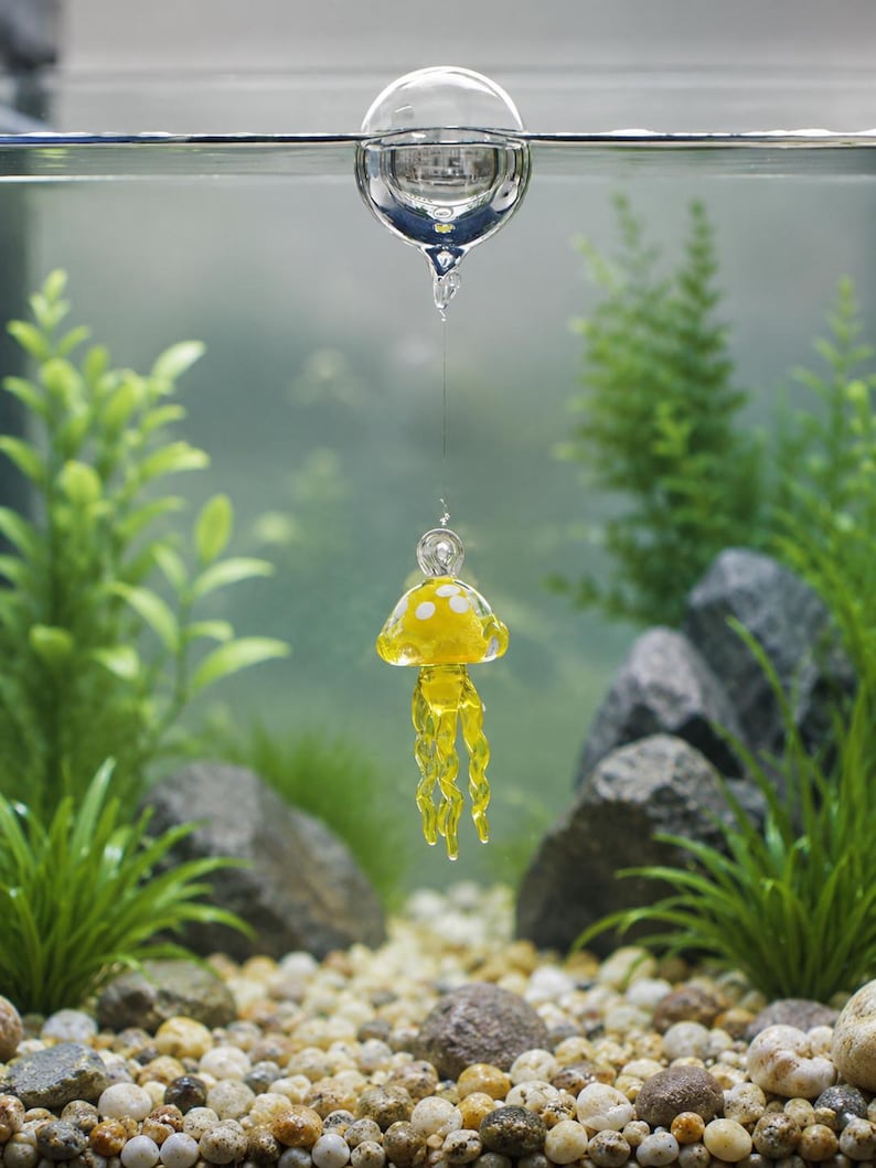 May include: A clear glass sphere floats at the water surface, connected by a thin filament to a small, yellow glass jellyfish ornament. The jellyfish features a rounded bell with white spots and several dangling, twisted glass tentacles.