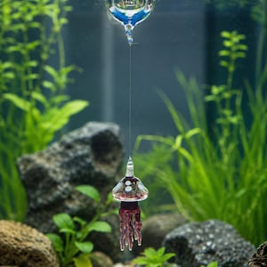 May include: A clear glass sphere floats at the water surface, connected by a thin, transparent line to a small, bell-shaped glass jellyfish ornament. The jellyfish features a dark purple body with white spots and dangling, translucent purple tentacles.