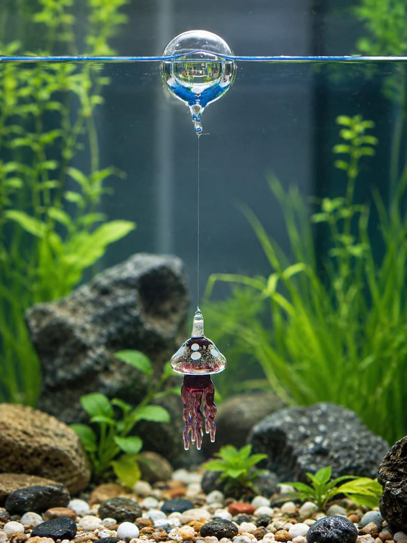 May include: A clear glass sphere floats at the water surface, connected by a thin, transparent line to a small, bell-shaped glass jellyfish ornament. The jellyfish features a dark purple body with white spots and dangling, translucent purple tentacles.