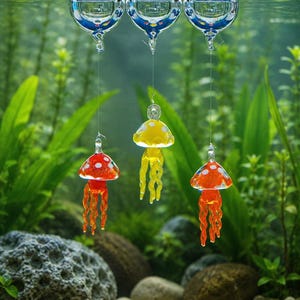 May include: Three glass jellyfish ornaments suspended from clear glass spheres floating at the water surface. The jellyfish are colored red, yellow, and orange, each featuring white spots on their caps and long, wavy, translucent tentacles.