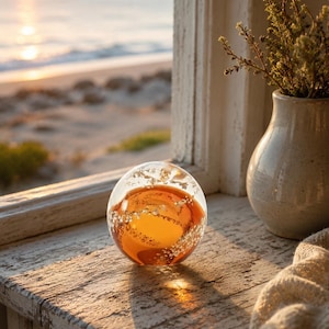 May include: A spherical glass paperweight featuring a deep amber-colored core surrounded by clear glass with embedded silver-toned metallic flakes. The object has a smooth, polished surface and reflects light through its translucent, rounded form.