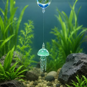 May include: A clear glass sphere floats on the water surface, connected by a thin, transparent line to a small, light green glass jellyfish ornament with white spots and dangling tentacles suspended beneath the surface of the water.
