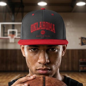 Può includere: Un cappellino da basket Oklahoma nero e rosso con la scritta "OKLAHOMA" ad arco sul davanti in lettere rosse. Il cappellino ha una visiera rossa e una grafica di pallacanestro. Le parole "Western Pride" sono sotto.