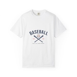 Op de afbeelding: Een wit t-shirt met korte mouwen en een afbeelding op de borst met de tekst BASEBALL in marineblauw, een honkbal, twee gekruiste marineblauwe knuppels, de rode tekst EST. 197 en de marineblauwe tekst BUILT BY HARD WORK.